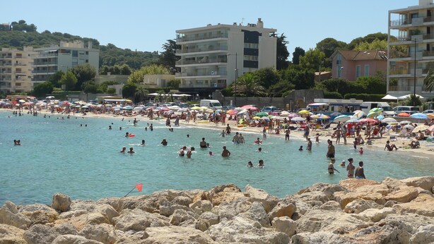 Une plage bondée où l’on voit des gens nager et bronzer, entourée de bâtiments et de parasols.