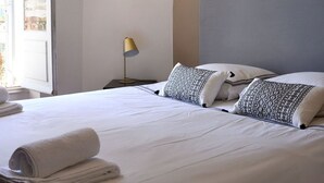 Un lit bien fait, avec du linge de lit blanc et des coussins à motifs, un miroir rond accroché au mur et une lampe posée sur une table de chevet.