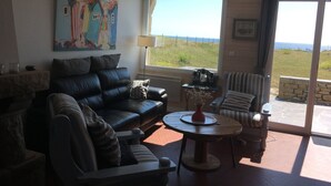 A living room with a black leather sofa, a striped armchair, and a round wooden coffee table. A painting is on the wall, and there is a view of the outdoors through a large window.
