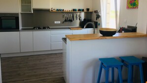 Une cuisine moderne avec des armoires blanches, un comptoir en bois et une cuisinière intégrée. On y trouve deux tabourets bleus près de l’îlot central, et une fenêtre avec des rideaux sur le côté droit.