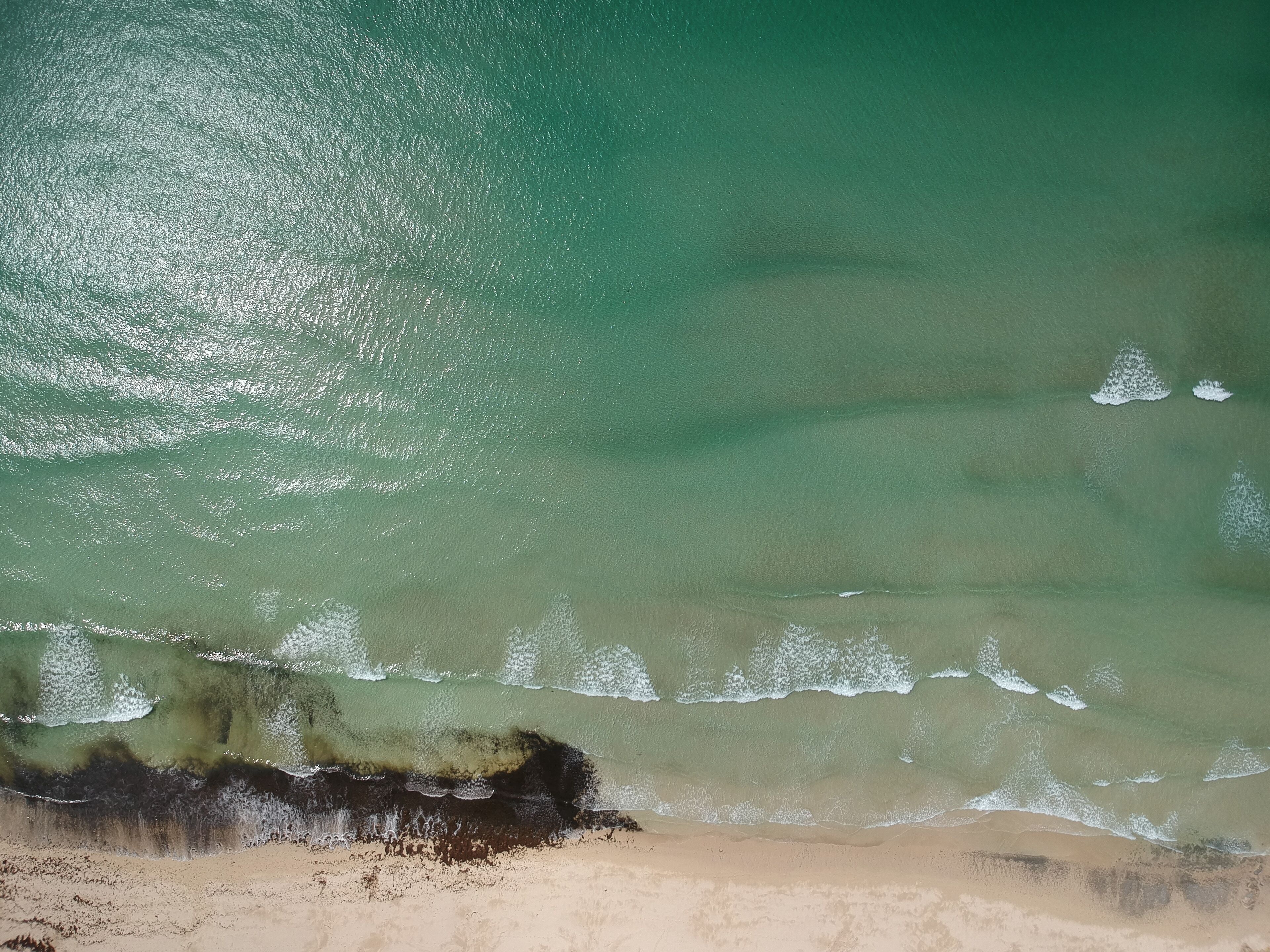 Une vue aérienne d’une plage aux eaux turquoise et limpides, avec de douces vagues et un rivage sablonneux.