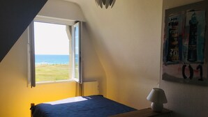 Une chambre avec un lit, une table de chevet, une lampe, une fenêtre donnant sur la plage et un mur orné d’une affiche.