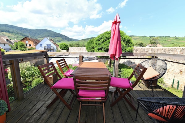 A wooden deck with a table and chairs, a pink umbrella, and a view of a green landscape with houses in the background.