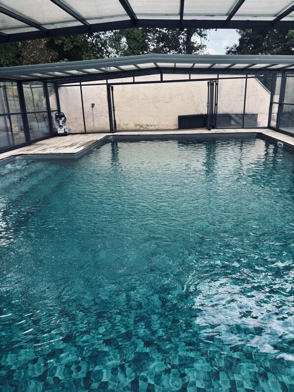 Una piscina interior cristalina con un deck de madera, rodeada de paredes de vidrio y un techo de metal.