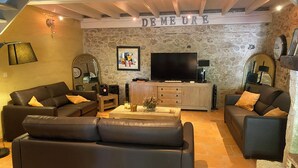 A living room with two leather sofas, a coffee table, a TV, and a stone wall with the word "DEMEURE" above it.