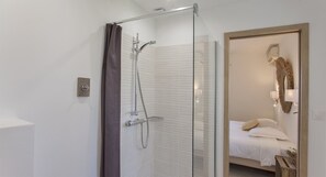 Une salle de bain moderne dotée d’une cabine de douche en verre, d’un espace de douche carrelé de blanc et d’un lit avec une tête de lit en bois dans la pièce adjacente.
