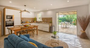 A modern kitchen with wooden cabinets, a dining area with a blue sofa, and a view of the garden through a glass door. - Villa "Albizzia" with terrace and garden 6 to 8 people (Calvi)