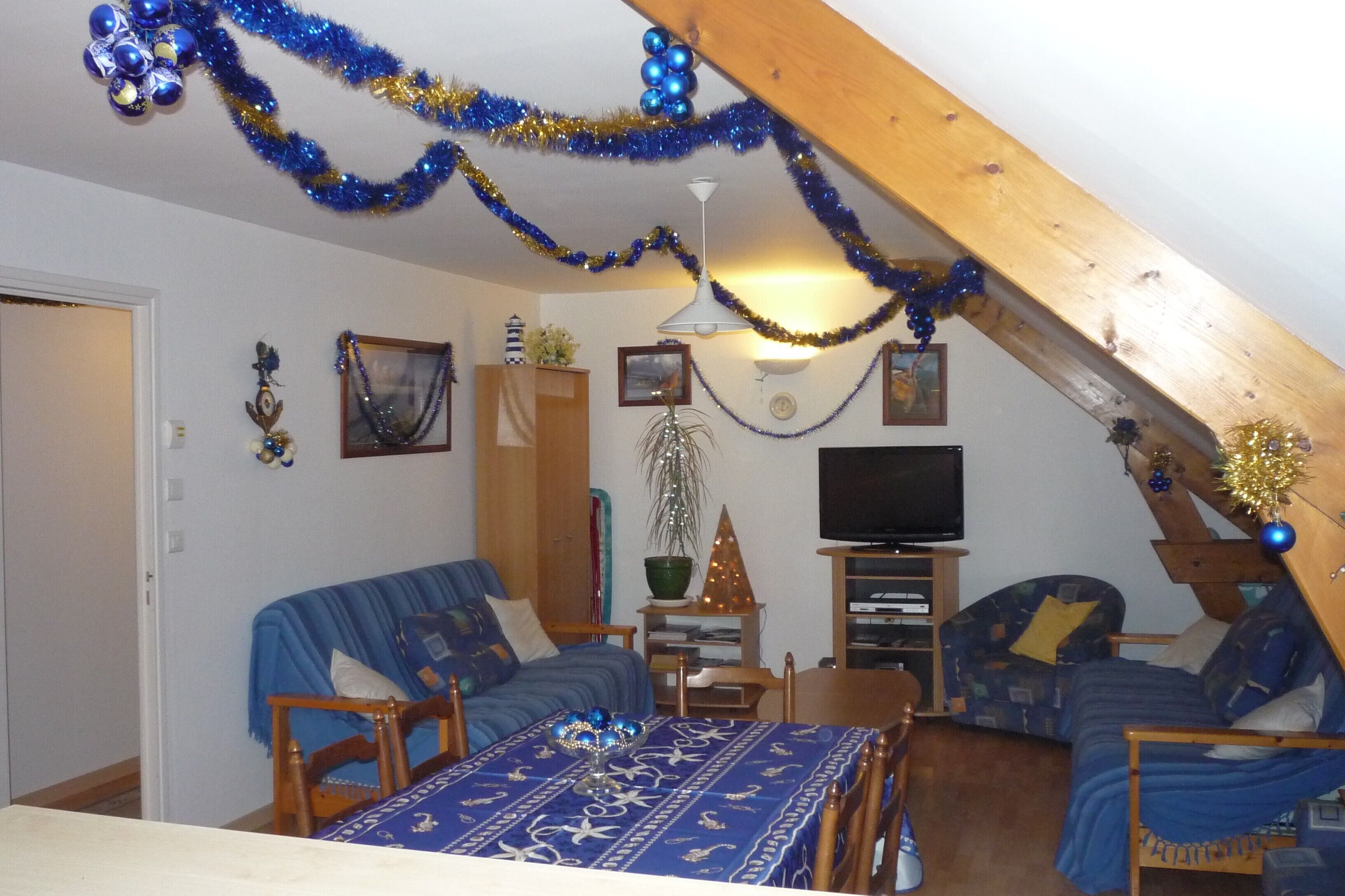 Une pièce mansardée chaleureuse avec un plafond en pente, des poutres en bois, une table à manger recouverte d’une nappe bleue, deux canapés bleus, un téléviseur et des décorations festives aux couleurs bleu et or.