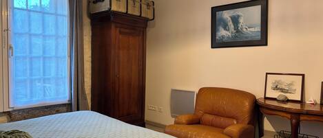 Un dormitorio con cama, un armario de madera, un sillón de cuero, una mesita de noche con un marco de fotos y una ventana con cortinas.