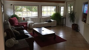 Een woonkamer met een witte bank, een rode fauteuil, een bruine bank, een houten salontafel en een groot raam met uitzicht op groen.