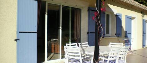 Une terrasse extérieure avec des chaises blanches et une table, une porte-fenêtre coulissante et des volets bleus sur le bâtiment.