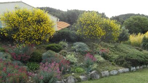 Un jardín ajardinado con diversos arbustos y plantas en flor, un muro de piedra y un edificio con techo de tejas al fondo.