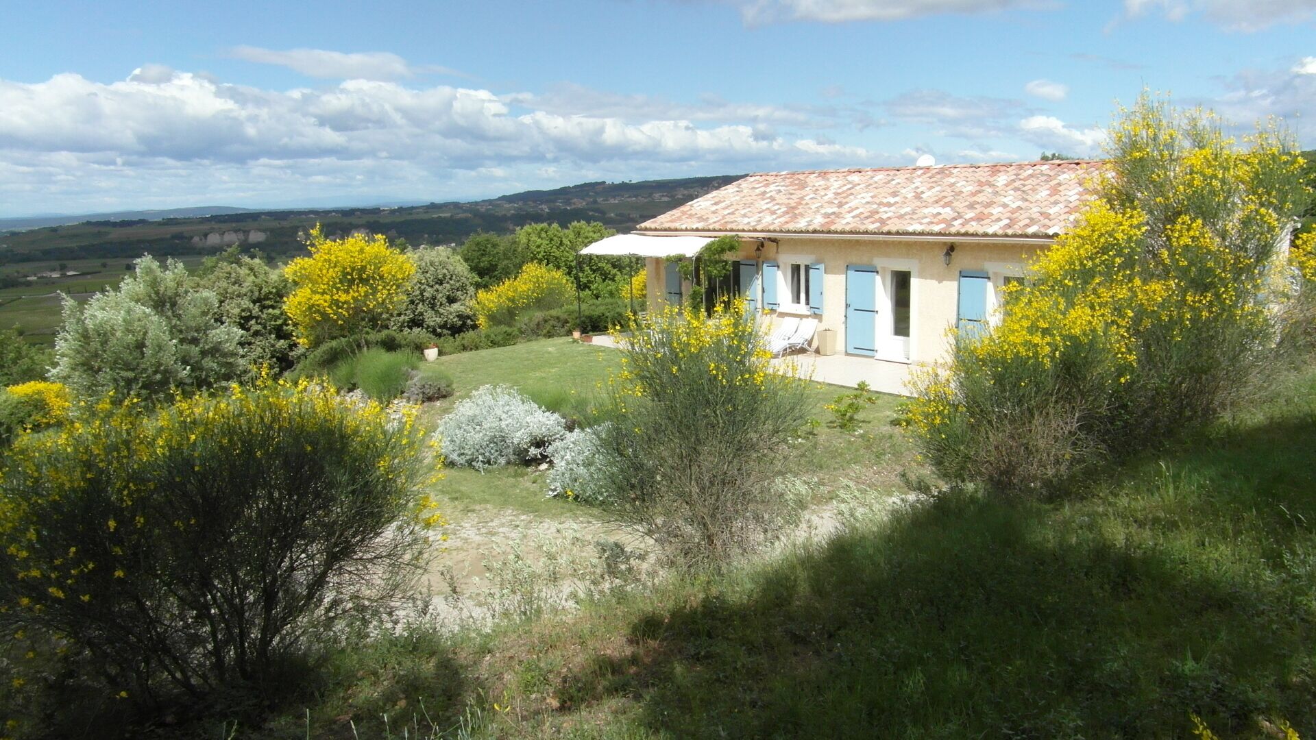 Une maison avec un toit en tuiles, entourée de verdure et de plantes à fleurs jaunes.