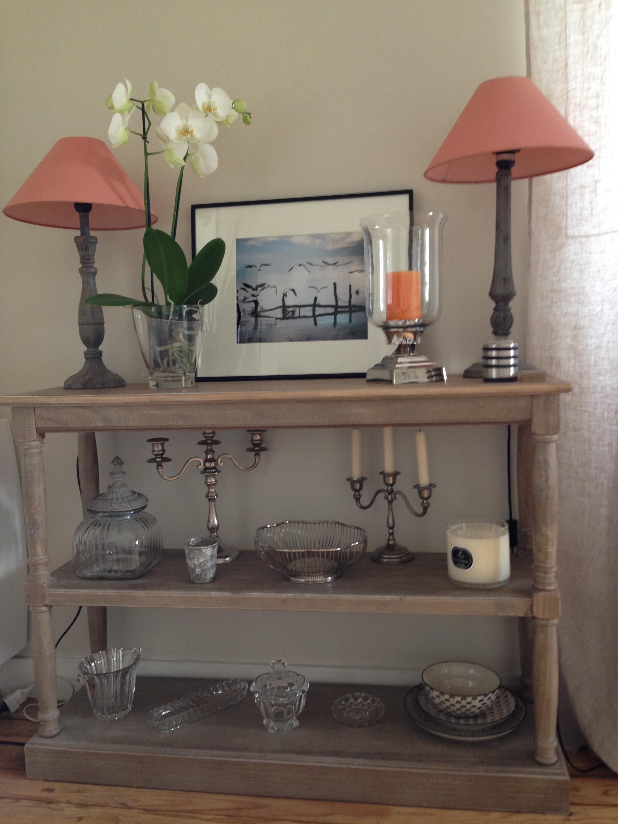 A wooden console table with a framed picture, two lamps, a vase with flowers, and various glassware and candles.