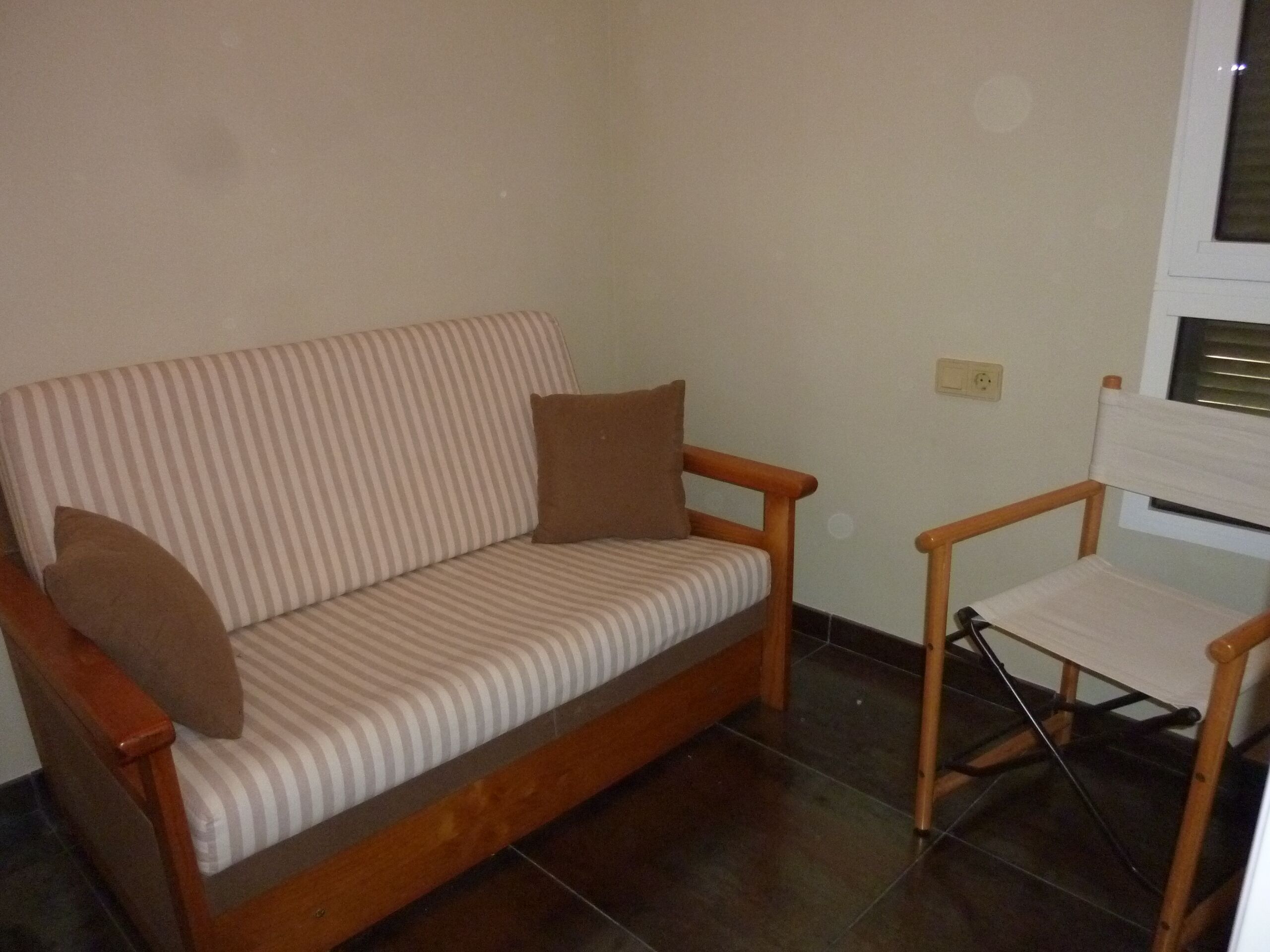 A wooden sofa with striped cushions and a matching armchair in a room with tiled flooring and a plain wall.