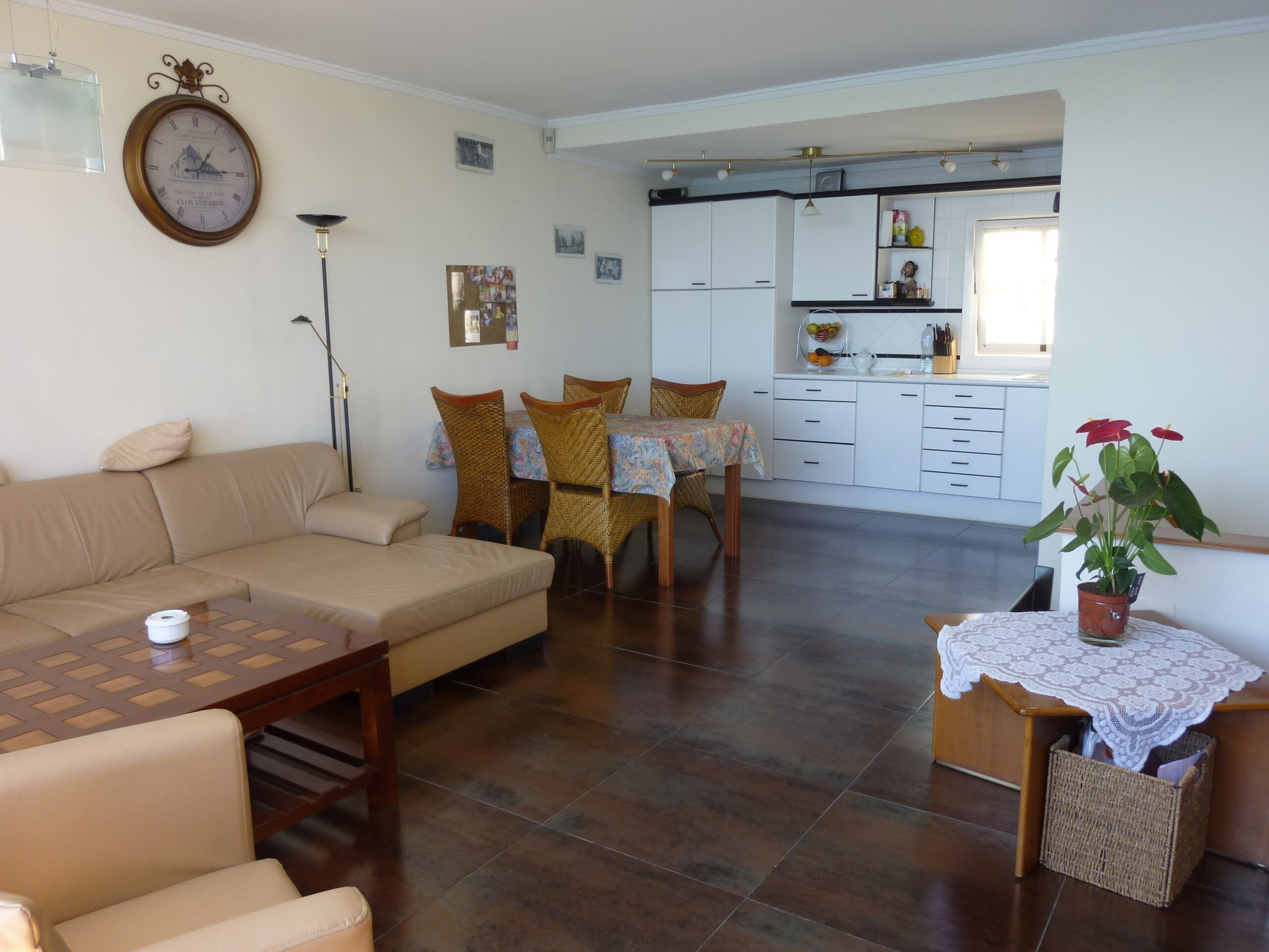 A living room with a sofa, a dining table with chairs, a kitchen area with cabinets, and a clock on the wall.
