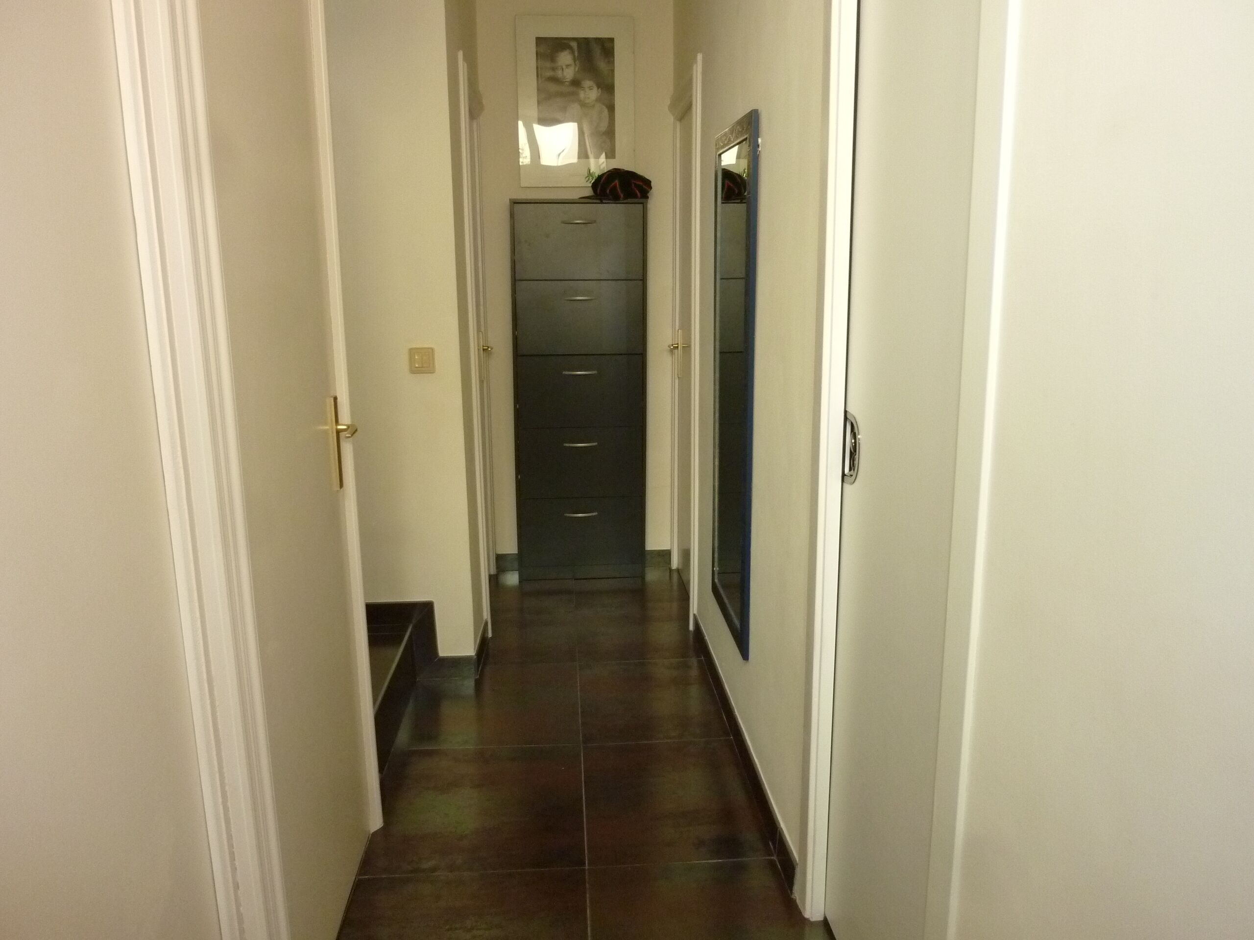 A narrow hallway with a dark tiled floor, white walls, and a door on the left.