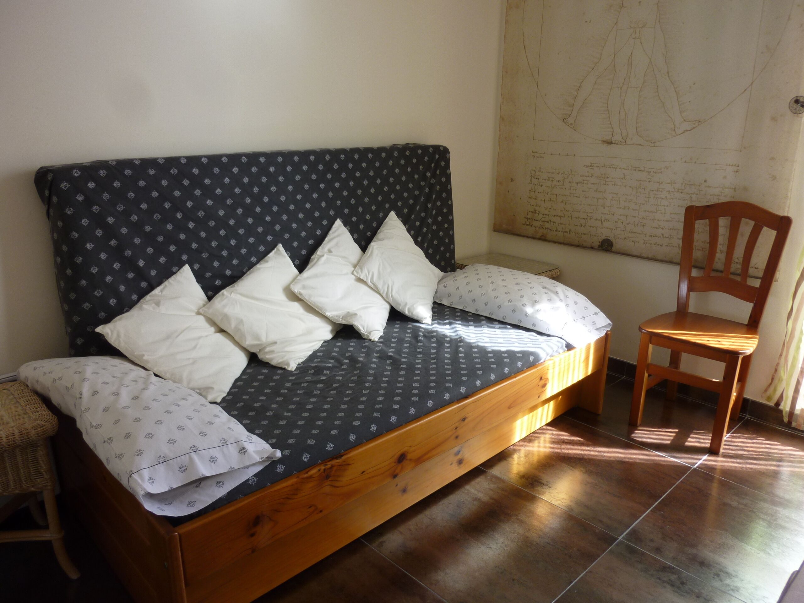 A single bed with a patterned headboard, white pillows, and a wooden frame. A wooden chair is positioned to the right of the bed.