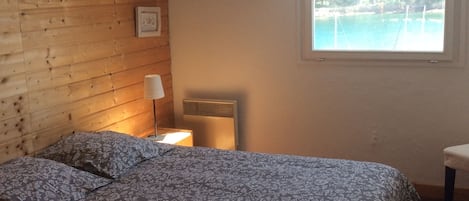 Ett sovrum med ett träpanelat tak, en säng med blommigt sänglinne, ett nattduksbord med en lampa och ett fönster med utsikt över en strand.
