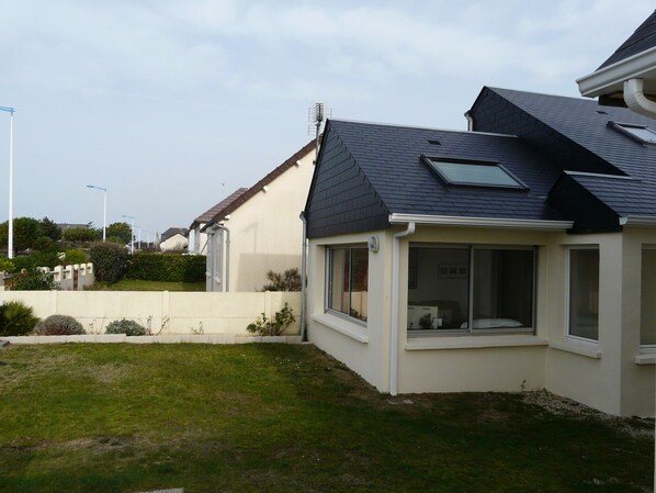 Une maison d’habitation avec un toit en pente, un petit garage attenant et un jardin devant la maison, agrémenté d’un petit muret et de quelques plantes.
