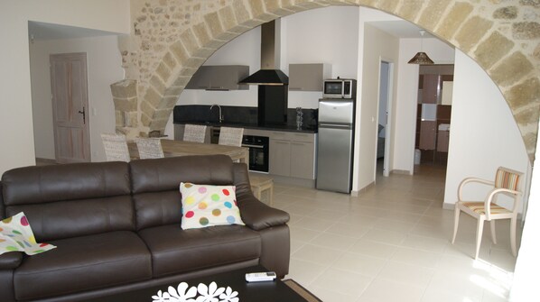 A living room with a brown sofa, a small table, and a polka-dotted pillow. There is a kitchen area with a stove, microwave, and refrigerator. An archway leads to another room. - Character cottage situated in the old center of a village for 4 people (Montagnac)