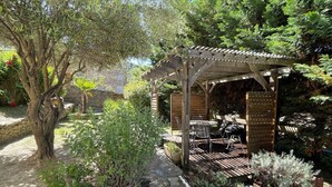 Een houten pergola met een tafel en stoelen, omringd door verschillende planten en een kiezelpad.