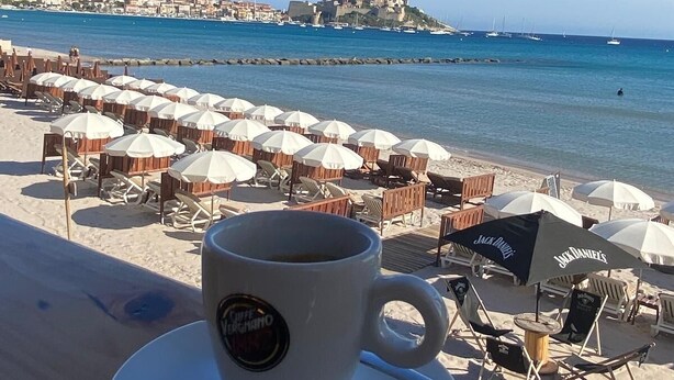 Een café aan het strand met uitzicht op zee en een kasteel in de verte.