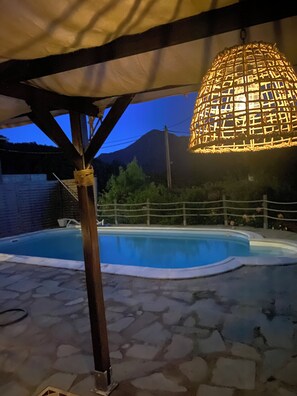 A swimming pool with a pergola overhead, a hanging light fixture, and a mountainous backdrop.