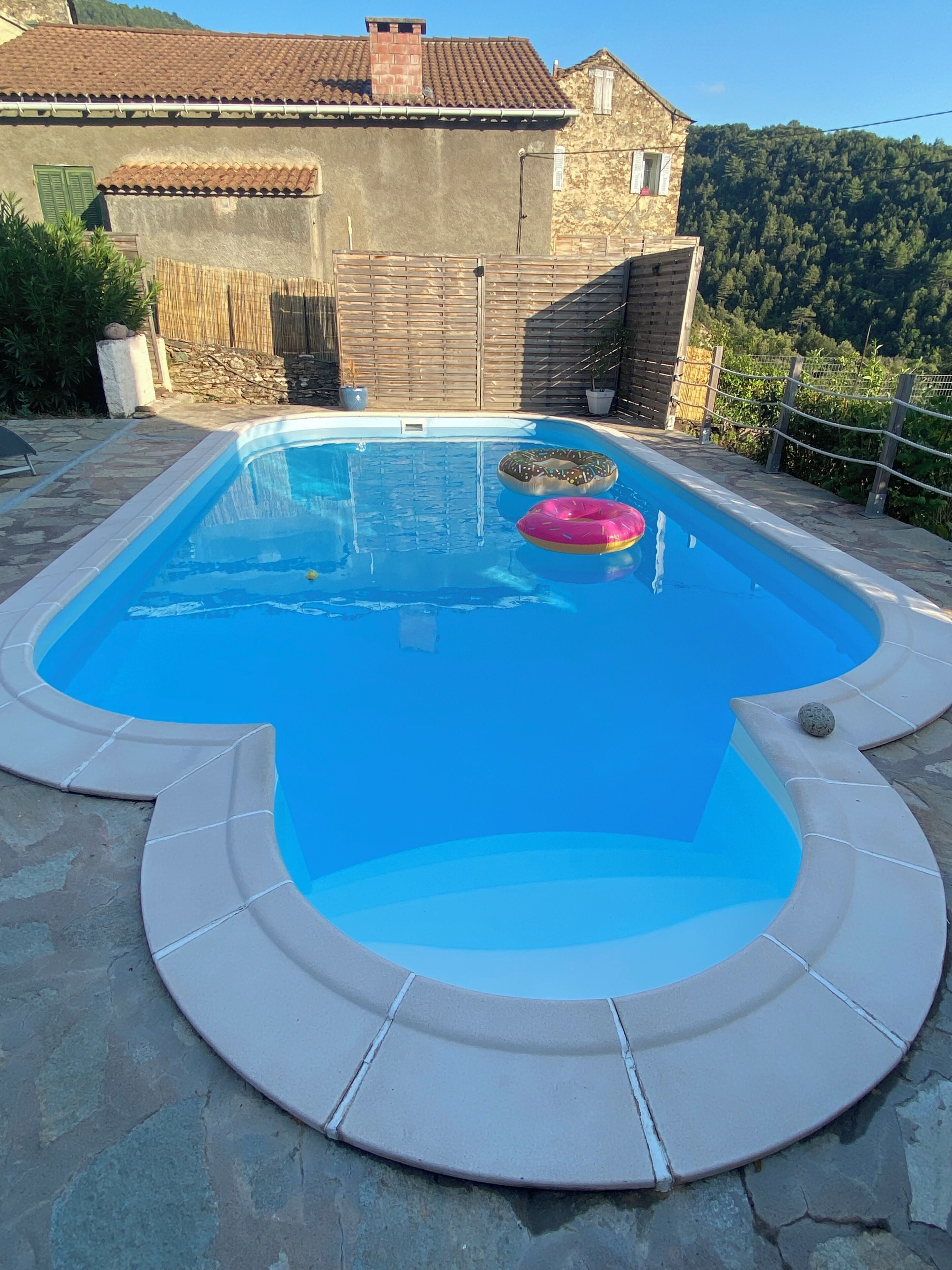 Una piscina con un deck de piedra, una cerca de madera y una casa al fondo.