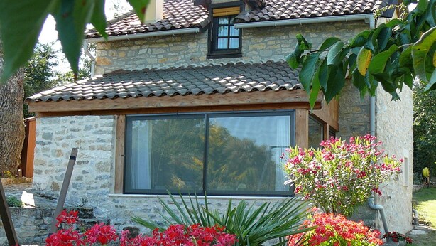 Uma casa de pedra com telhado de telhas, uma janela grande e um jardim com flores vermelhas.