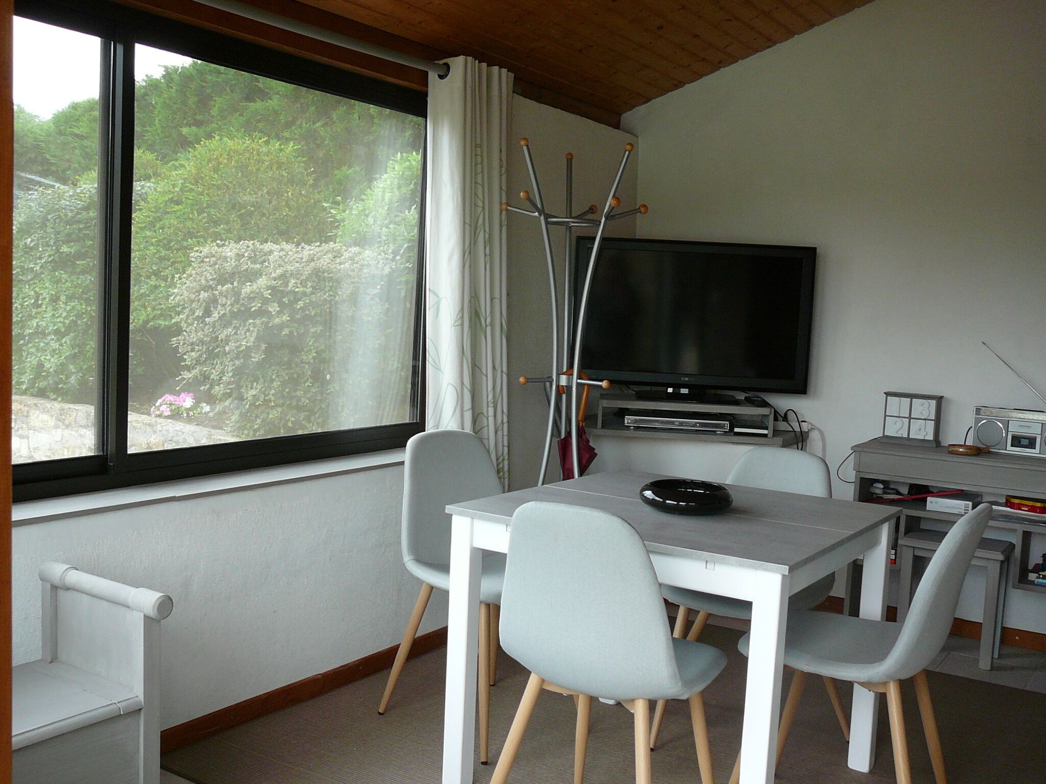 A room with a TV, a table with chairs, a window with curtains, and a view of greenery outside.
