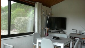Quarto com televisão, mesa e cadeiras, janela com cortinas e vista para o verde lá fora.