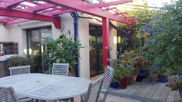 Un patio con mesa y sillas, rodeado de plantas en macetas y una pérgola.