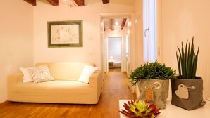 A living room with a beige sofa, a wooden floor, a framed artwork on the wall, and two potted plants.