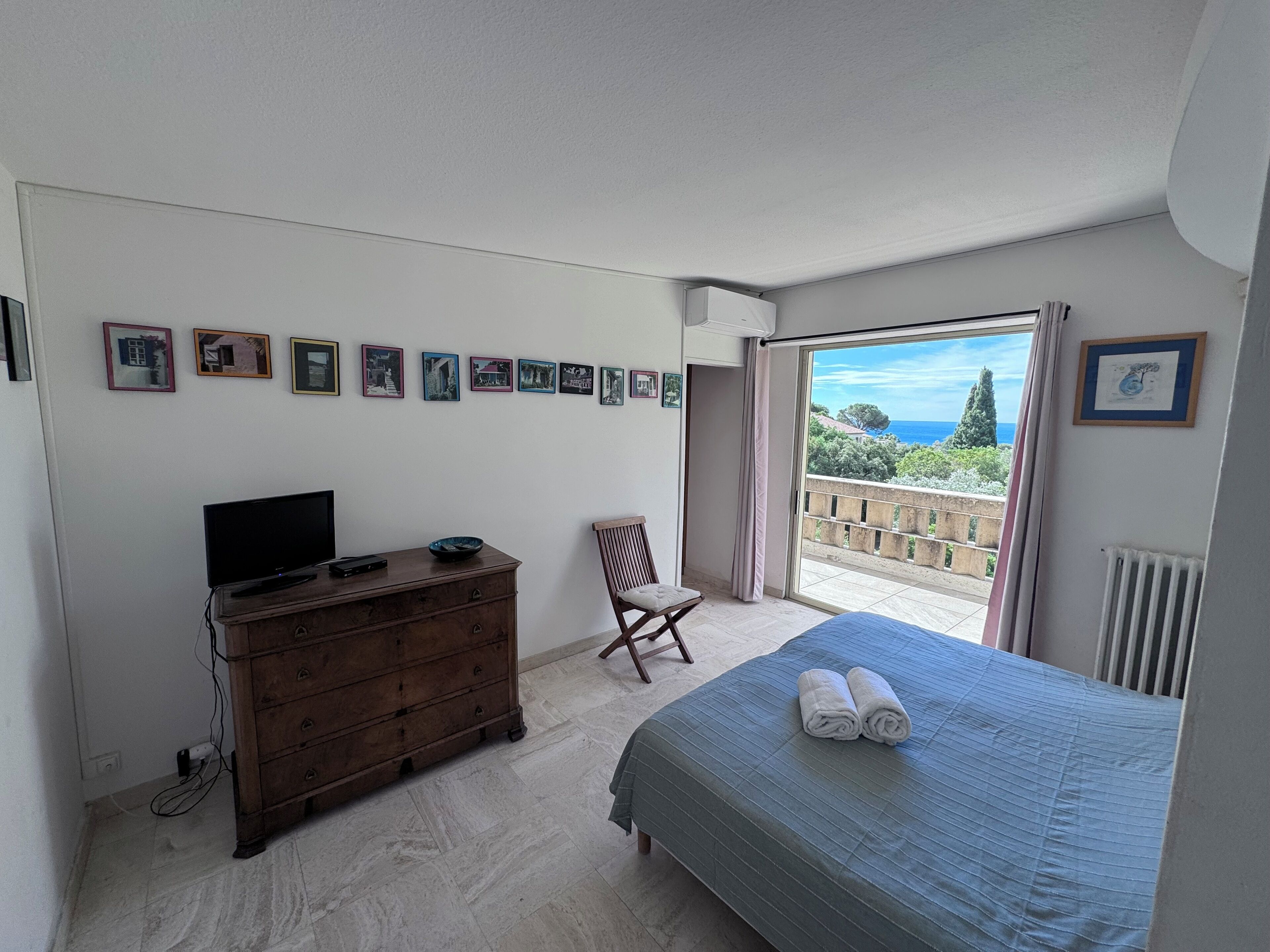 A bedroom with a bed, a dresser, a chair, and a TV. There are pictures on the wall and a view of the sea through the window.