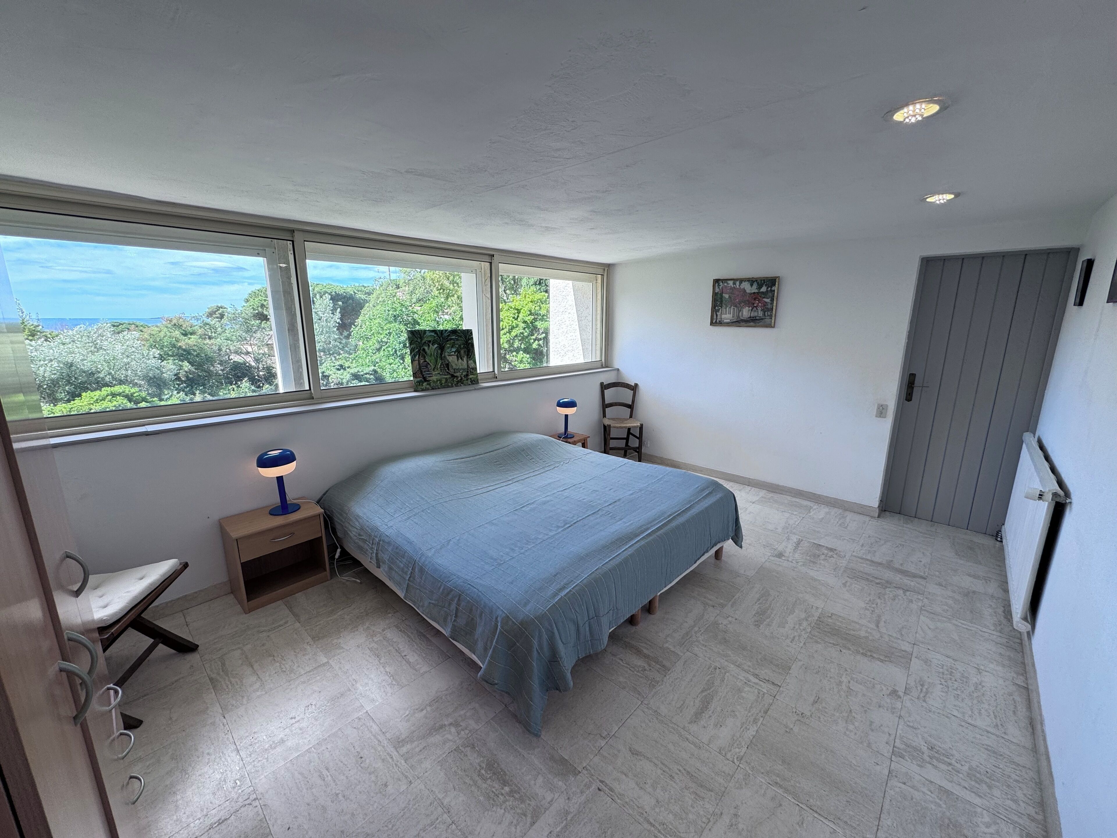 A bedroom with a bed, a desk, a chair, a window with a view, and a door.