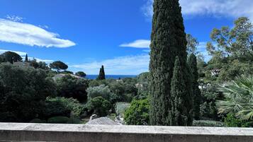 Une vue depuis un balcon orné d’une balustrade en pierre, donnant sur un jardin planté de divers arbres et offrant une vue lointaine sur la mer.