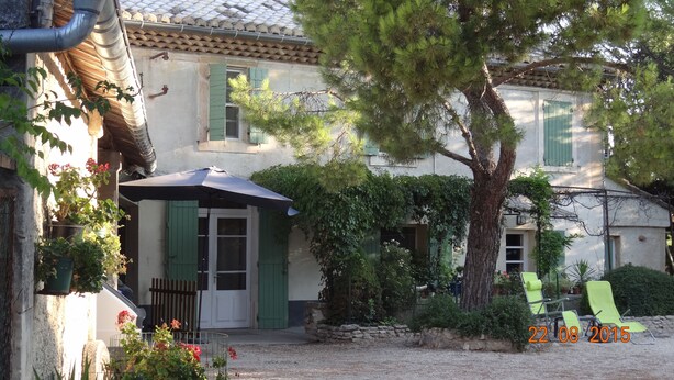 Il s’agit d’un bâtiment traditionnel, doté d’un toit en tuiles, d’une entrée couverte et d’un mur en pierre. Les fenêtres sont ornées de volets verts, un arbre se trouve dans le jardin, et une date est inscrite dans un coin.