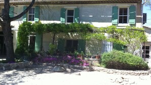 Un bâtiment à deux étages avec des volets verts, un toit en tuiles et un jardin où poussent des fleurs violettes.