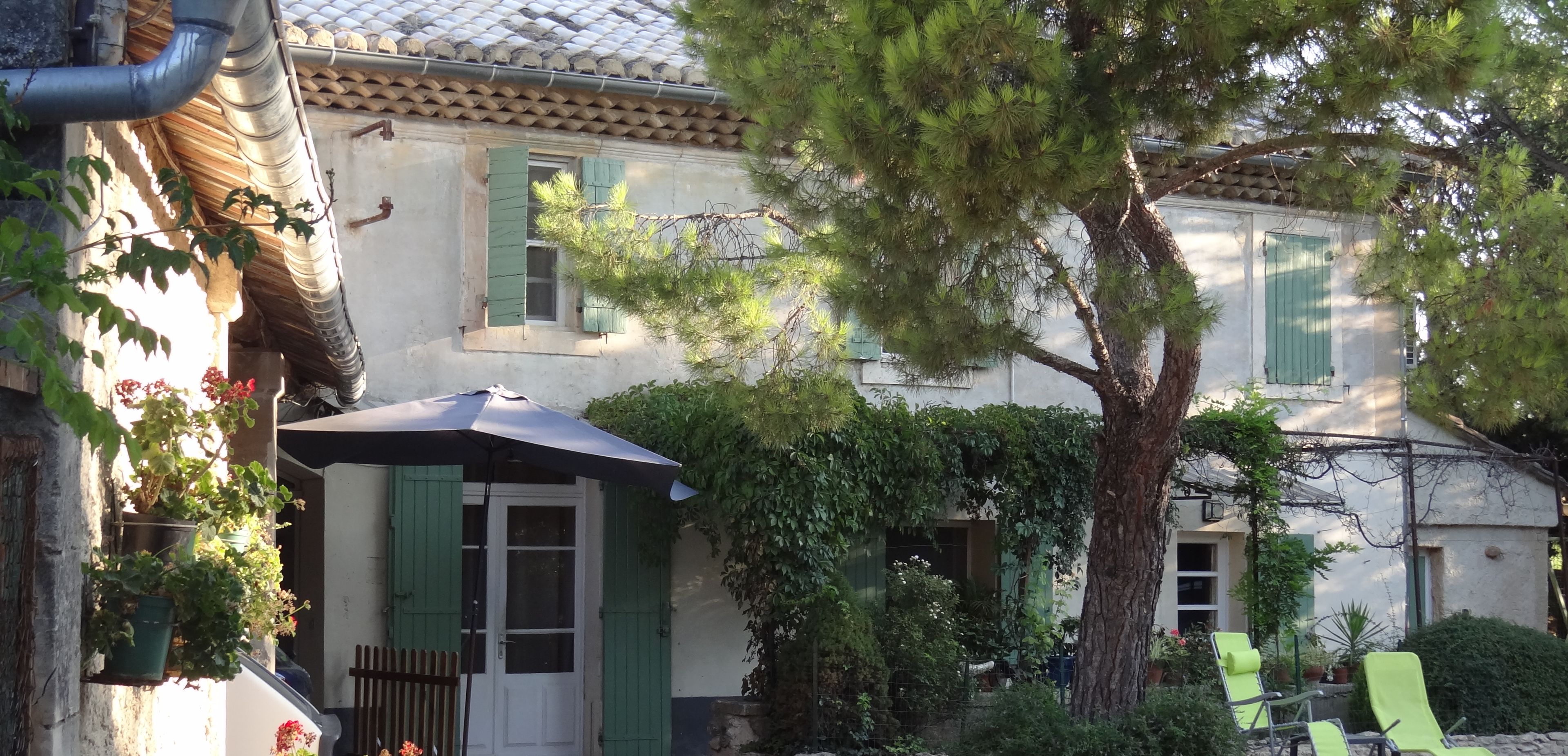 A traditional building with a tiled roof, a covered entrance, and a stone wall. There are green shutters on the windows, a tree in the front yard, and a date stamp in the corner.