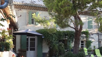 A traditional building with a tiled roof, a covered entrance, and a stone wall. There are green shutters on the windows, a tree in the front yard, and a date stamp in the corner.