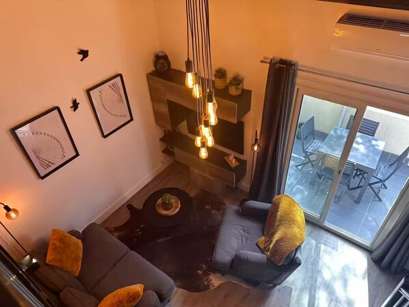 A modern living room with a sofa, a coffee table, and a chair. There are two framed artworks on the wall, a hanging light fixture, and a sliding glass door leading to an outdoor area.