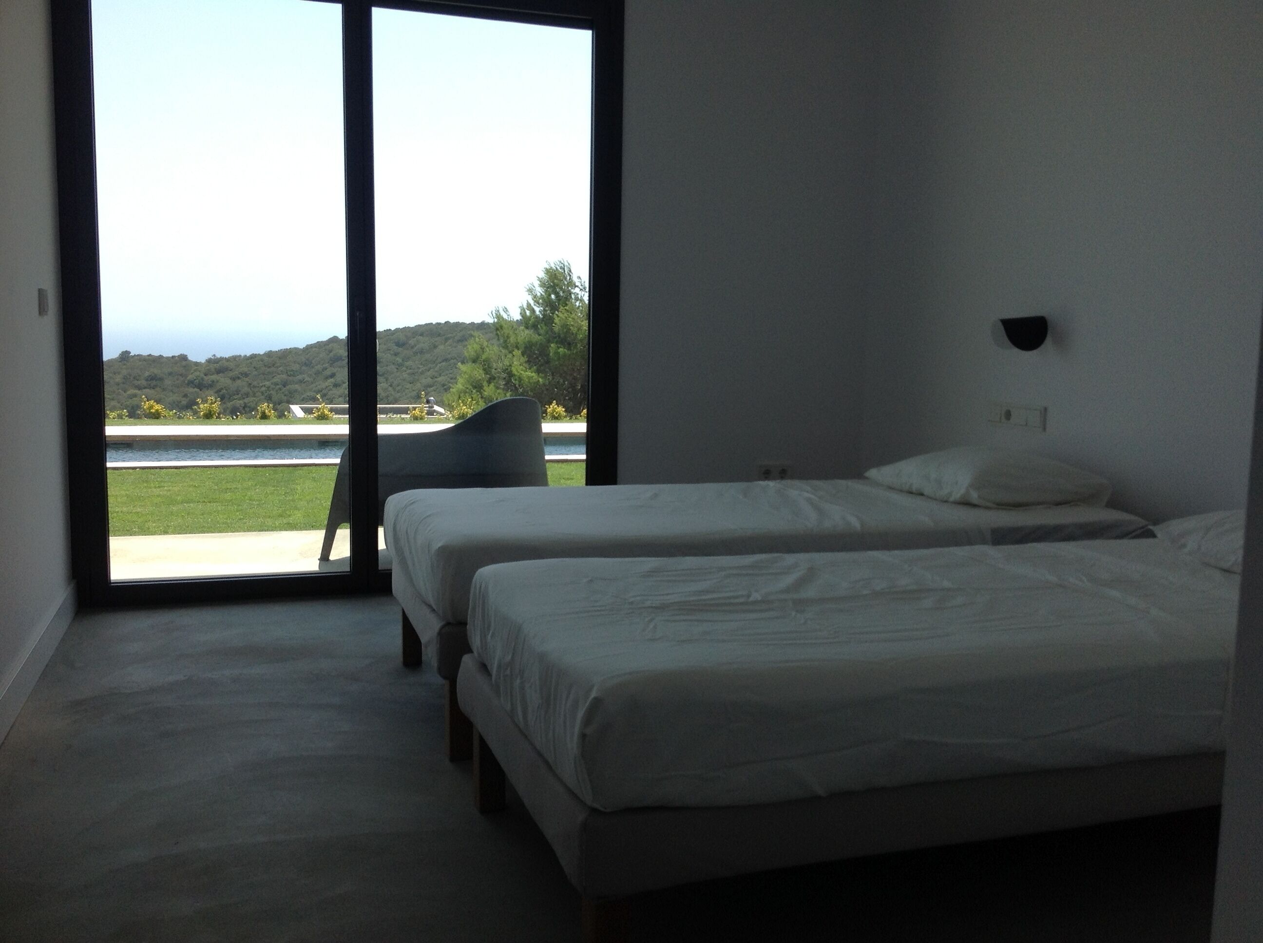 A bedroom with two beds, a chair, and a large window offering a view of a body of water and greenery.
