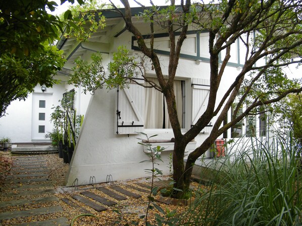 A white building with a sloped roof, a window with shutters, and a tree in front.