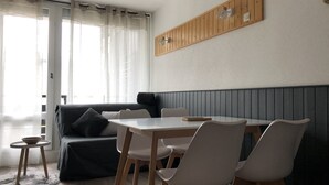 Uma sala de estar moderna com sofá, mesa de jantar e cadeiras. Há uma pequena mesa redonda com uma tigela sobre ela. O quarto possui uma parede de painéis de madeira e uma janela com cortinas leves.