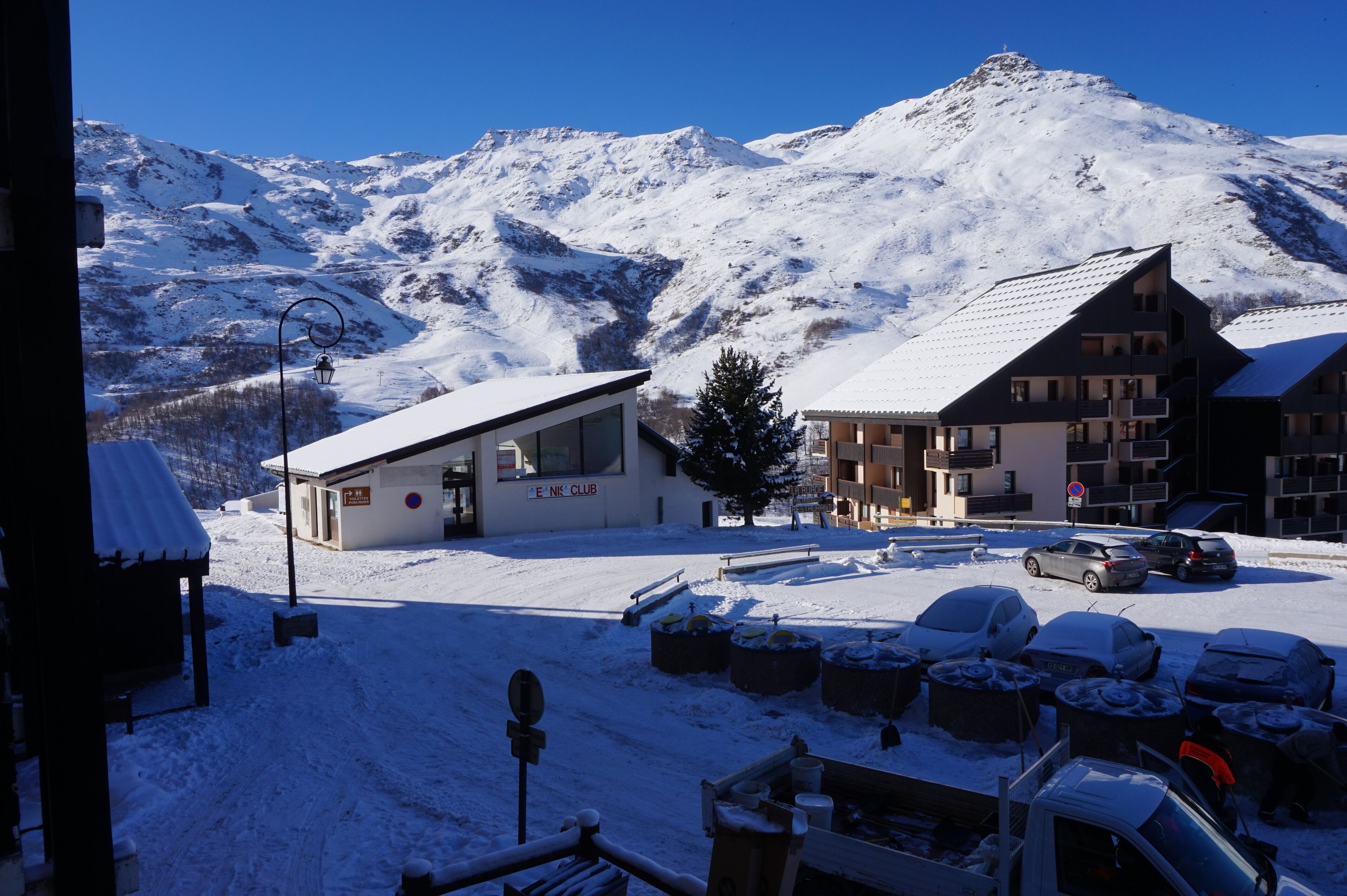 Een bergstad bedekt met sneeuw, met een gebouw van een skigebied, een restaurant en enkele geparkeerde auto's op de voorgrond.