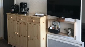 Um espaço compacto com armário de madeira, cafeteira, TV de tela plana e uma pequena prateleira com objetos decorativos.