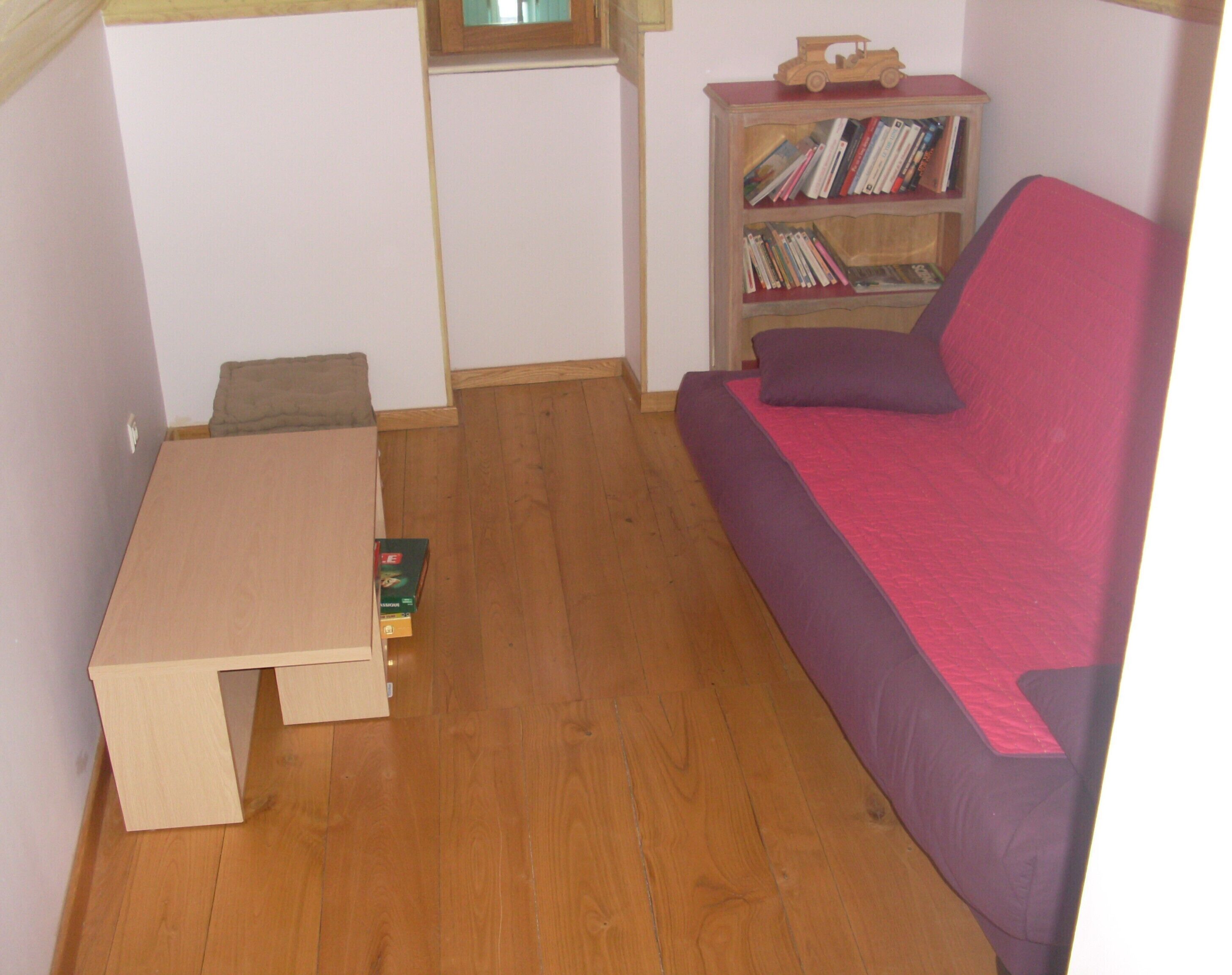 Una habitación pequeña con suelo de madera, un sofá púrpura, una mesita y una estantería llena de libros.