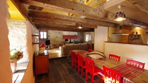 Un intérieur rustique avec une table à manger en bois, des chaises rouges et un coin cuisine équipé d’une cuisinière et de casseroles suspendues.