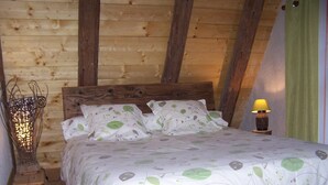 Un plafond en bois avec des poutres apparentes, un lit recouvert d’une couvre-lit à motifs de feuilles, une lampe de chevet et une décoration murale tissée.